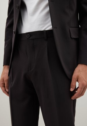 Veste de costume noire et pantalon ajusté ; le tissu semble lisse ; deux boutons visibles au niveau de la taille ; t-shirt blanc en dessous.