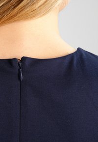 Närbild på en persons nacke och övre rygg med kort blont hår, iförd ett mörkblått plagg med en liten dragkedja bak.