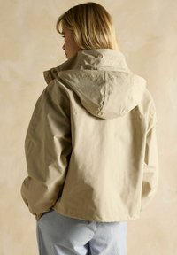 Jachetă bej cu capuciu, cu un finisaj mat, având un detaliu cu logo pe spate și o croială lejeră. Combinată cu pantaloni în dungi, oferă un aspect casual.