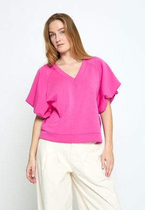 Kvinde med langt blondt hår iført en lys pink V-hals løs top og højtaljede, vidde hvide bukser, stående mod en ensfarvet baggrund.