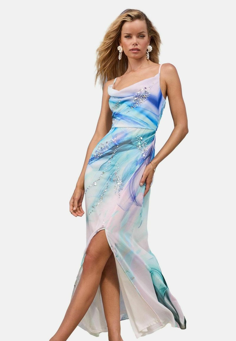 Femeie îmbrăcată într-o rochie vaporasă fără mâneci, cu modele abstracte albastre și purpurii și embellishments strălucitoare, pozând cu o picior vizibil printr-o fanta.