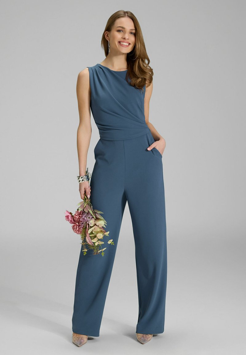 Een smaragdgroene, mouwloze jumpsuit met een gedrapeerd ontwerp, brede pijpen en zakken, gecombineerd met een bloembouquet en puntige hakken.