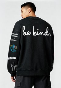 Črni bombažni pulover s tekstom "bodi prijazen." na hrbtni strani, pisana grafika na rokavu in dodatni tekstovni detajli. Rebrasti manšetni in rob.