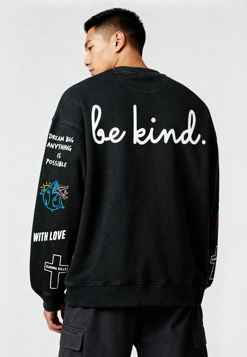Črni bombažni pulover s tekstom "bodi prijazen." na hrbtni strani, pisana grafika na rokavu in dodatni tekstovni detajli. Rebrasti manšetni in rob.