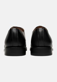 Brogue in pelle nera con finitura liscia, punta rotonda e tacco rettangolare basso. L'interno presenta una fodera marrone a contrasto.