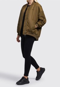 Personne portant une veste bomber surdimensionnée vert olive, legging noir et baskets noires, debout avec une main dans la poche sur un fond uni.