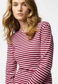 Junge Frau mit welligem blondem Haar, die ein rot-weiß gestreiftes langärmeliges Oberteil vor einem einfarbigen weißen Hintergrund trägt.