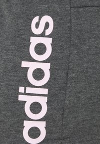 Pantalones de chándal grises con una textura suave, que presentan un destacado logo "adidas" en rosa impreso verticalmente a lo largo del costado. Bolsillo visible.