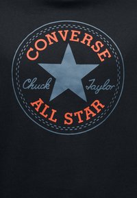 Μαύρο βαμβακερό μπλουζάκι με μεγάλο κυκλικό σχέδιο που απεικονίζει ένα μπλε αστέρι και κόκκινο κείμενο με τις λέξεις "CONVERSE", "Chuck Taylor" και "ALL STAR".
