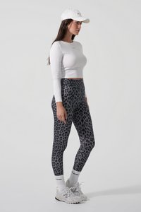 Graue Leggings mit Leopardenmuster, eng anliegendes Design. Weißes cropped Langarmshirt. Weiße Mütze. Helle Sport-Schuhe mit schwarzen Akzenten.