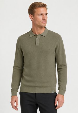 Man draagt een olijfgroene geribbelde poloshirt met lange mouwen en zwarte broek, kijkt naar zijn rechterkant tegen een effen lichte achtergrond.