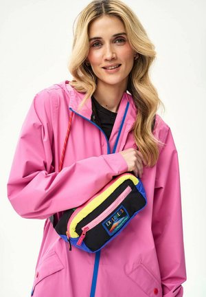 Blondynka w różowej kurtce przeciwdeszczowej trzyma czarną saszetkę na pasku z żółtymi, niebieskimi i różowymi elementami oraz napisem „EXPLORERS”.