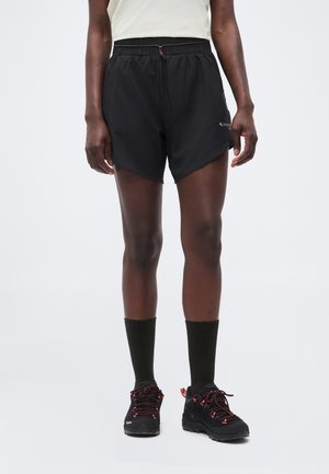 Személy fekete sportnadrágban, fekete, középmagas szárú zokniban és piros fűzős fekete futócipőben fehér háttér előtt.