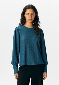Teal tröja med en texturerad tyg som har diamantmönster, puffärmar och rund halsringning. Matchad med svarta byxor.