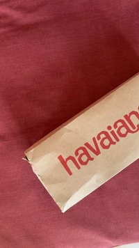Rudas kartoninė dėžutė su raudonu "havaianas" tekstu, gulinti ant bordo spalvos audinio paviršiaus. Dėžutėje matomi nedideli įlenkimai ir atvira kraštas.