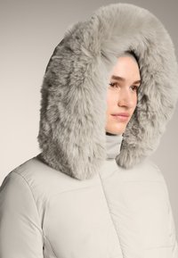Femeie îmbrăcată într-o geacă de iarnă gri deschis, cu o glugă mare și pufoasă din blană, poziționată în lateral, pe un fundal simplu.