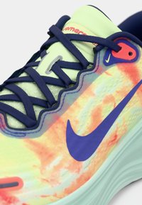 Αθλητικό παπούτσι Nike με πολύχρωμο επάνω μέρος σε πορτοκαλί, κίτρινες και πράσινες αποχρώσεις, μπλε κορδόνια και μοβ λογότυπο Swoosh. Υφή με ανάγλυφη επιφάνεια.