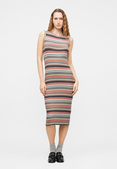 Femme debout portant une robe en maille sans manches, de longueur au genou, à rayures horizontales rose, noire, grise et beige, associée à des chaussettes grises et des mocassins noirs.