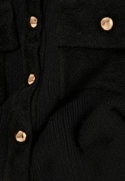 Tissu noir texturé avec de grands boutons ronds dorés brillants disposés verticalement et une petite poche carrée en haut à droite.