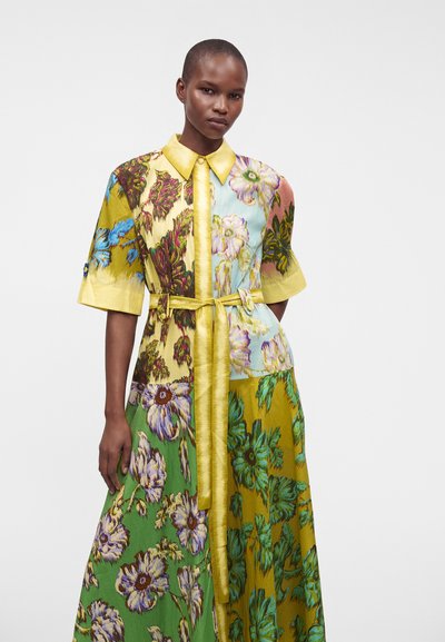 Femme aux cheveux courts portant une robe fleurie multicolore avec un col et une ceinture jaunes, debout devant un fond blanc uni.