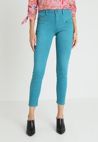 Tyrkysové skinny džíny vyrobené z pružné látky, se zapínacími předními kapsami a elegantním designem, doplněné černými vysokými podpatky.