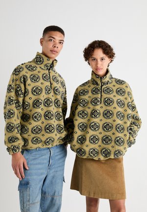 Fleece zip-up jackets in yellow with blue floral patterns, featuring high collars and elastic cuffs, paired with denim and corduroy bottoms.