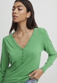 Grünes langärmliges Oberteil mit V-Ausschnitt, das eine Knopfleiste und strukturiertes Strickmuster aufweist. Weicher Stoff mit figurbetontem Design.