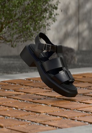 Czarna skórzana sandałka z grubą podeszwą i paskiem na sprzączkę, unosząca się nad drewnianymi kafelkami z szarymi kamyczkami na zewnątrz, blisko zielonego krzewu.