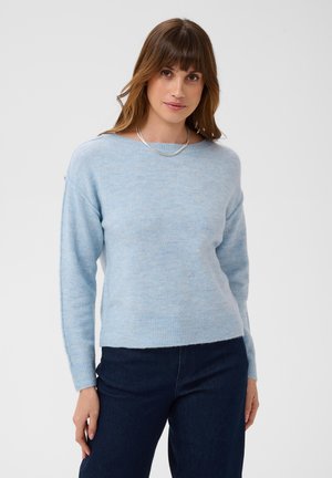 Vrouw met bruin haar die een lichtblauwe trui met lange mouwen en donkerblauwe spijkerbroek draagt, staand tegen een effen lichte achtergrond.