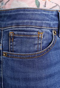 Jeans de mezclilla de color azul desgastado, con un bolsillo trasero con costuras naranjas y un acento de botón de metal. La textura parece suave.