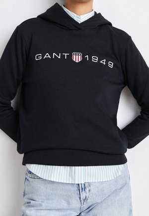Person trägt einen schwarzen GANT Hoodie mit „GANT 1949“ und einem Schild-Logo, darüber ein blau-weiß gestreiftes Hemd und hellblaue Jeans.