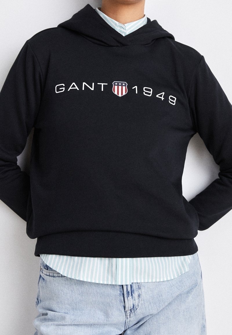 Personne portant un sweat à capuche noir GANT avec « GANT 1949 » et un logo bouclier, superposé à une chemise rayée bleue et blanche et un jean bleu clair.