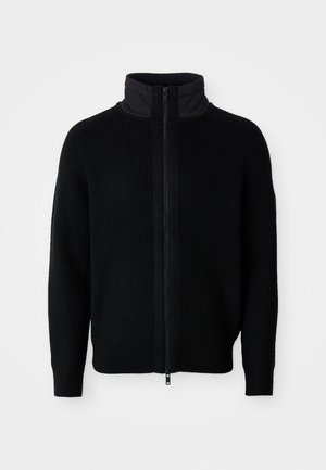 Sweater preto com um colarinho alto, fecho com zípper, textura canelada e mangas longas. O tecido combina painéis de malha e lisos.
