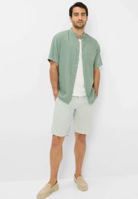 Lichtgroen korter t-shirt met knopen en een kraag, gecombineerd met lichtgepatenteerde shorts en beige instappers. Casual outfit ontwerp.