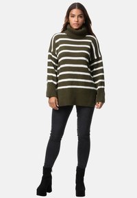 Grüner Strickpullover in Oliv mit weißen Streifen, featuring einen hohen Rollkragen, überschnittene Schultern und einen lockeren Schnitt. Getragen mit schwarzen Skinny Jeans.