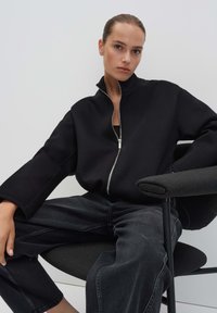 Zwarte zip-up jack met een hoge kraag, met brede mouwen, in combinatie met donkere spijkerbroeken. Model zittend in een stoel tegen een lichte achtergrond.