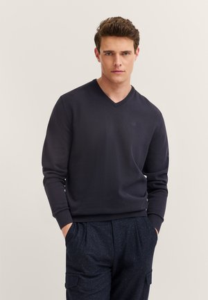 Junger Mann mit kurzen braunen Haaren, der einen dunkelblauen V-Ausschnittpullover und gesprenkelte marineblaue Cargo-Hosen trägt, steht mit den Händen in den Taschen.