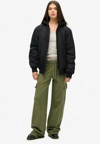 Veste bomber noire, débardeur gris ajusté, pantalon cargo olivâtre surdimensionné avec poches, et chaussures noires. Tissu lisse, coupe décontractée.