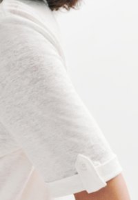 Camisa blanca de manga corta con un acabado texturizado, que presenta un puño enrollado con un detalle de botón. Material ligero y ligeramente transparente.