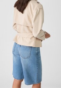 Beige jacket with double-breasted design, side pockets, and a tied waist, paired with light blue denim shorts featuring a classic cut and pockets.