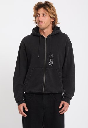 Jeune homme aux cheveux châtain clair portant un sweat à capuche noir zippé avec des lettres blanches verticales et un pantalon noir, regardant sur le côté.