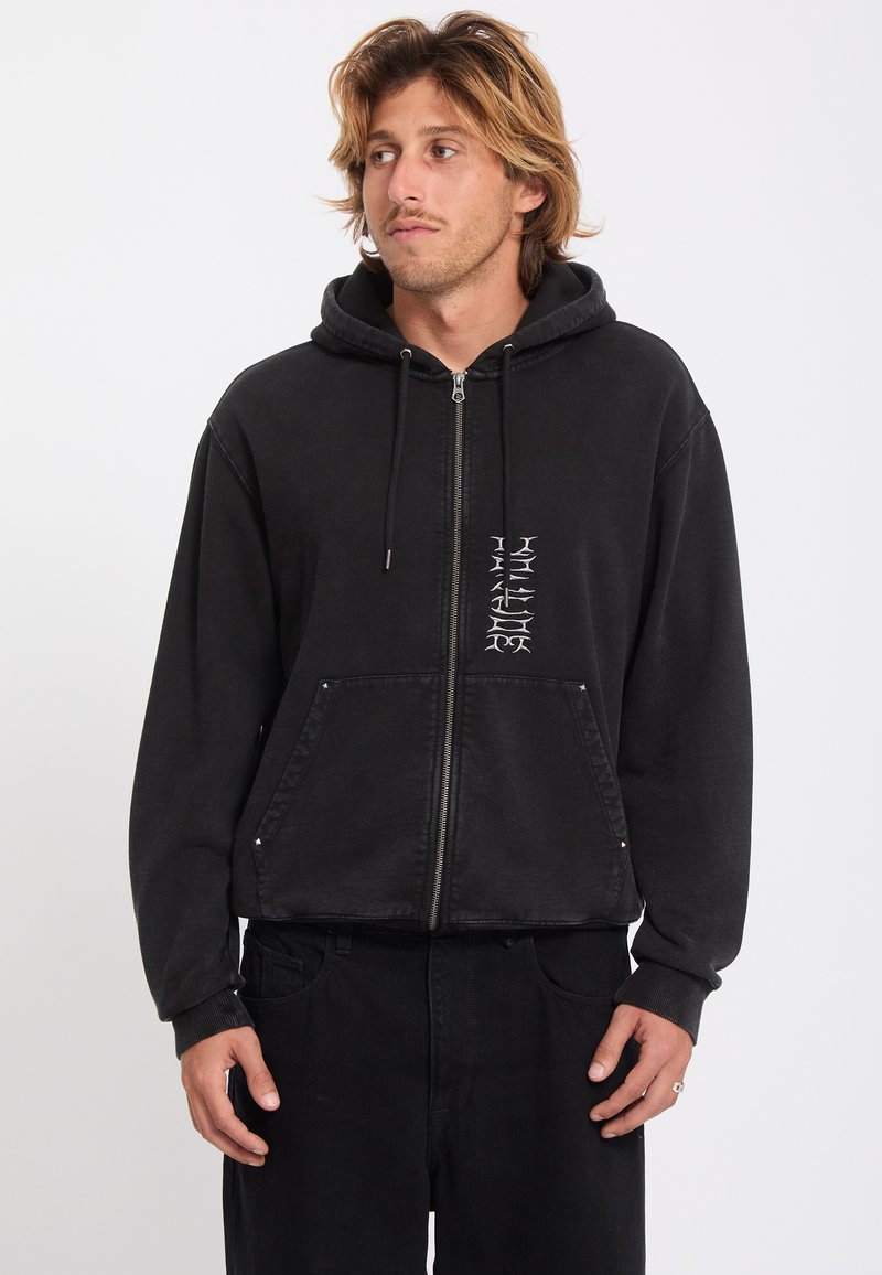 Jeune homme aux cheveux châtain clair portant un sweat à capuche noir zippé avec des lettres blanches verticales et un pantalon noir, regardant sur le côté.