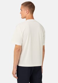 Vit t-shirt med kort ärm och rund halsringning, med en mjuk textur, sedd bakifrån, tillsammans med mörka byxor. Inga synliga logotyper.