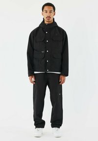 Giacca nera con molte tasche frontali e cappuccio, abbinata a pantaloni coordinati. Entrambi sono realizzati in un tessuto liscio e opaco, indossati con sneakers bianche.