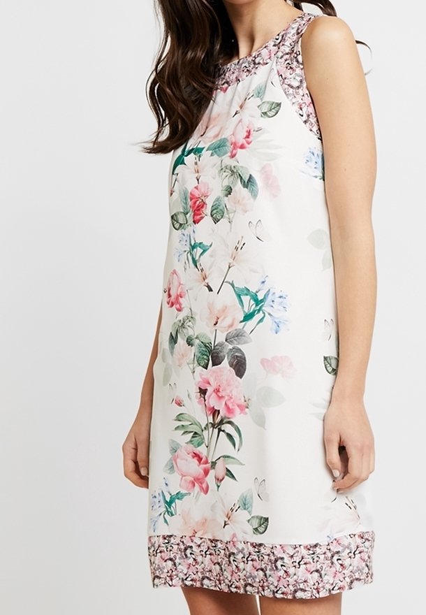 Robe blanche sans manches avec imprimé floral rose et vert, avec une bordure à motif à l'encolure et à l'ourlet, portée par une femme.