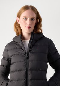 Black puffer jacket with a hood, featuring quilted segments, front zip closure, and a small logo on the chest. Smooth texture.
