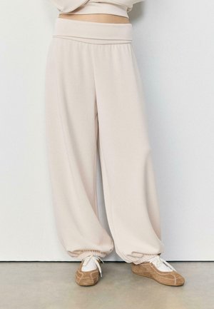 Beige Sweatpants mit weitem Bein und elastischen Bündchen, kombiniert mit weißen Socken und hellbraunen Mokassins aus Wildleder auf Betonboden.