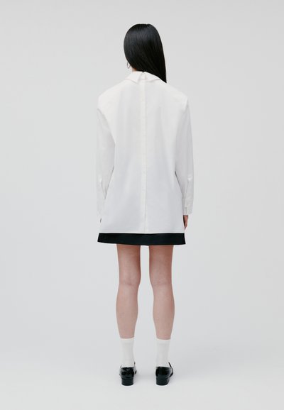 White long-sleeve shirt with back buttons, paired with a black skirt, worn over white socks and black shoes. Smooth fabric, straight fit.