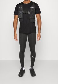 Mustapohjainen urheiluvesti, jossa on useita taskuja, mustan t-paidan päällä, yhdistettynä tiukkoihin mustiin legginsseihin ja mustiin lenkkikenkiin, joissa on valkoisia logo-aksentteja.