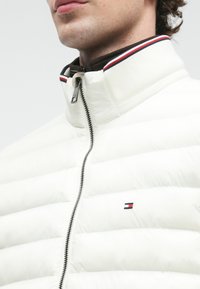 Vyras vilkintis baltą siūtu švarku su priekine užtrauktuku ir dryžuotu apykaklės detaliu, matoma dalis jo veido ir kaklo.